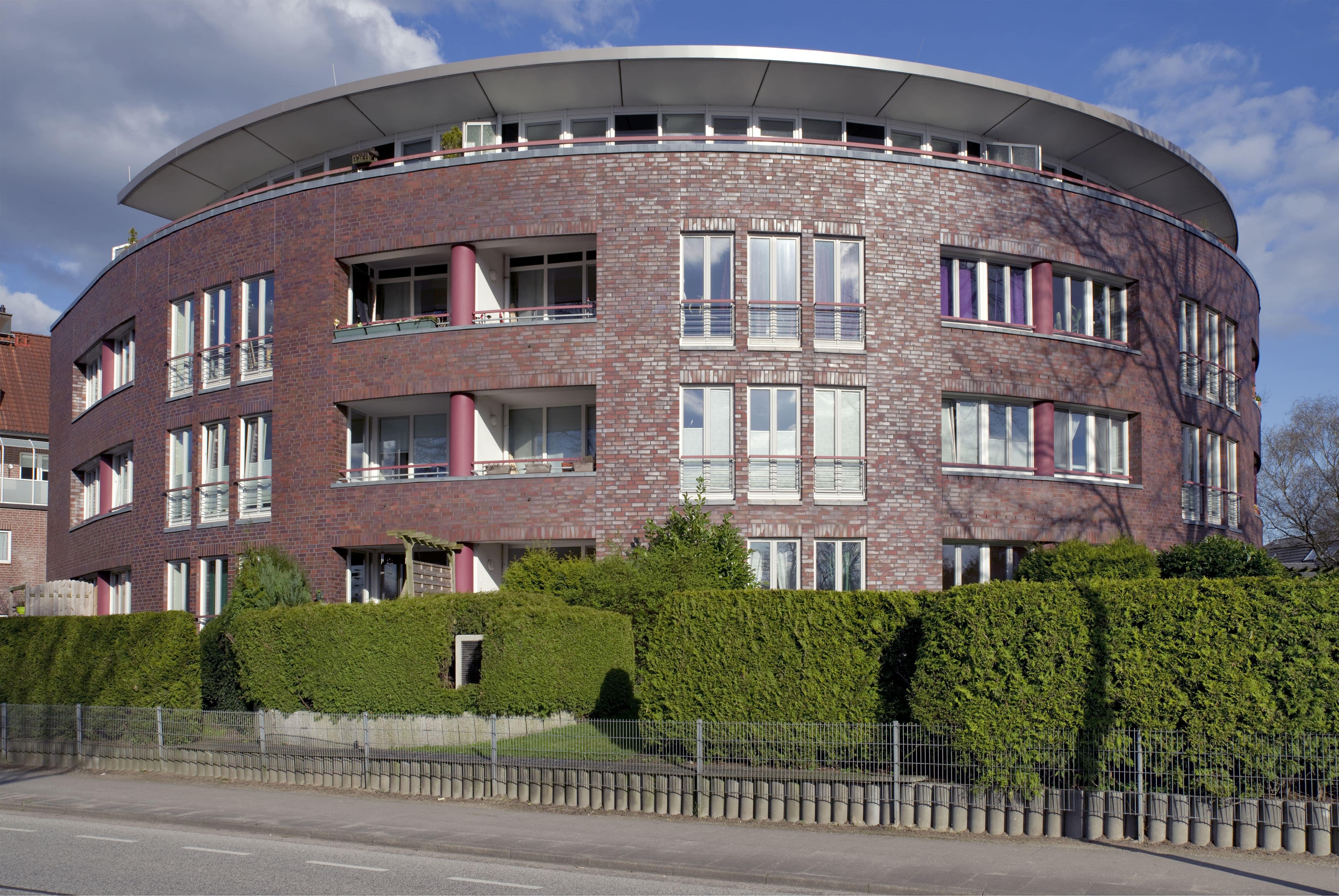 Ein modernes, rundes Mehrfamilienhaus aus rotem Klinker mit vielen Fenstern und Balkonen. Ein Beispiel für die Immobilien-Kapitalanlagen der Pensionskasse der Hamburger Hochbahn Aktiengesellschaft -VVaG-.