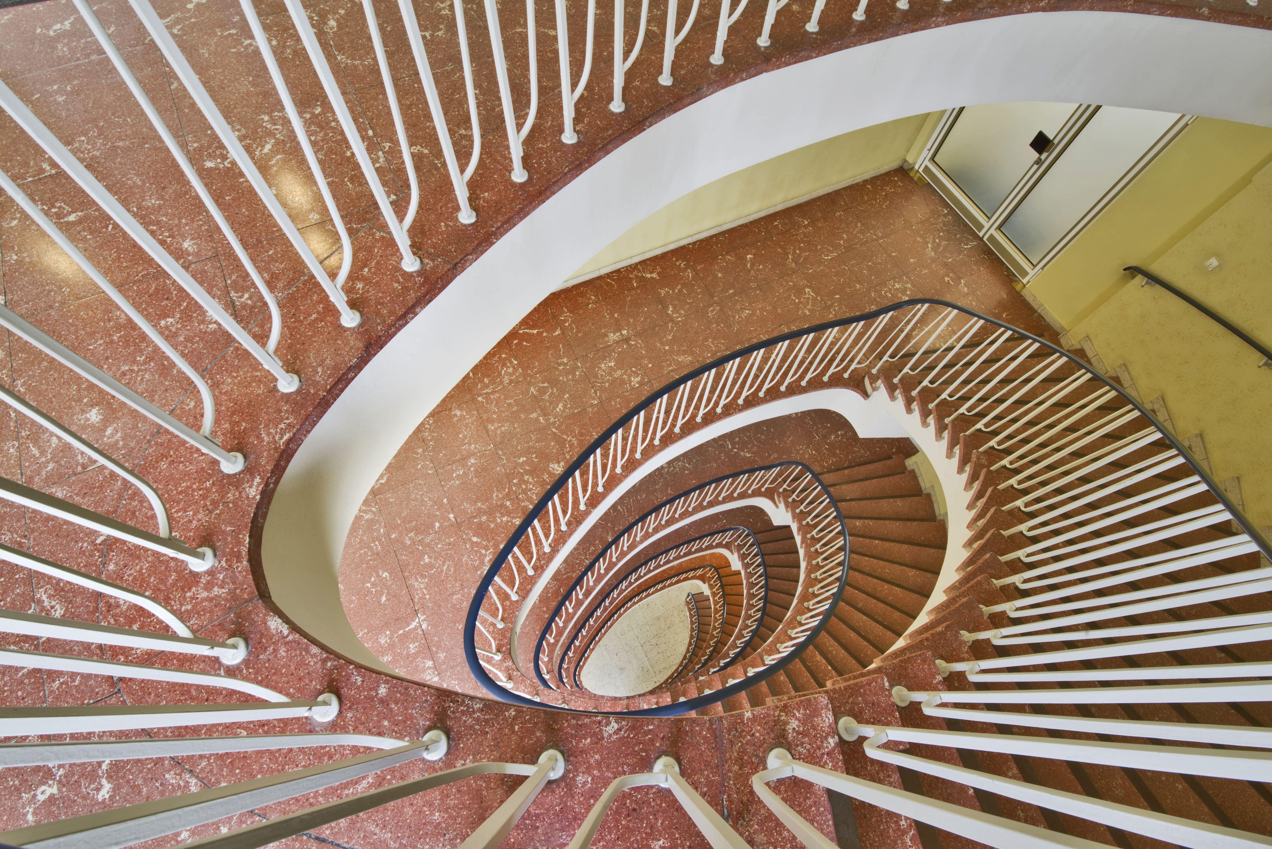 Eine von oben fotografierte, rote, geschwungene Wendeltreppe in einem historischen Gebäude. Das Bild symbolisiert den kontinuierlichen Aufstieg und die Stabilität der Pensionskasse der Hamburger Hochbahn Aktiengesellschaft -VVaG-.