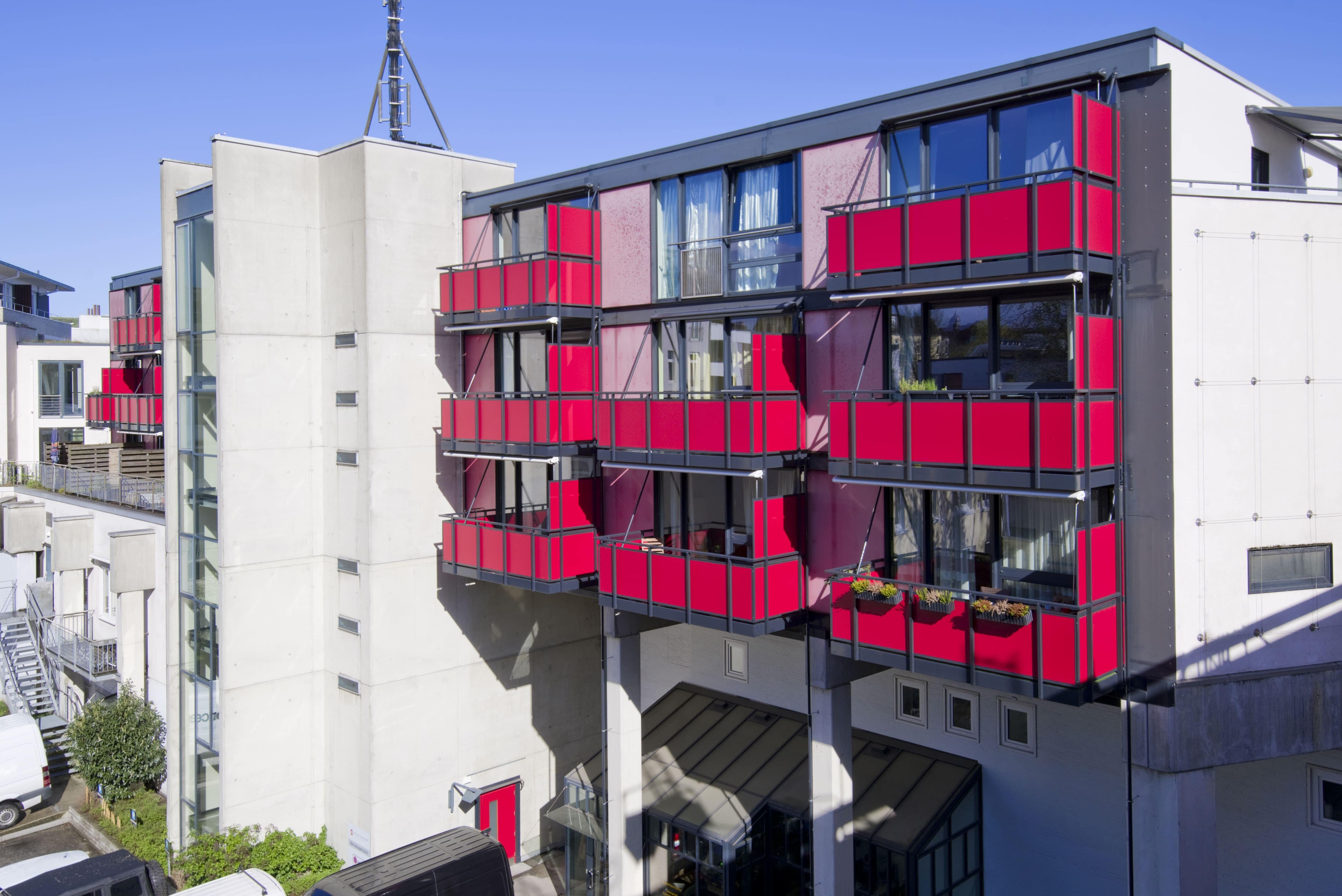Ein modernes, helles Mehrfamilienhaus mit auffälligen, leuchtend roten Balkonen. Das Gebäude ist ein Beispiel für die Immobilien-Kapitalanlagen der Pensionskasse der Hamburger Hochbahn Aktiengesellschaft -VVaG-.